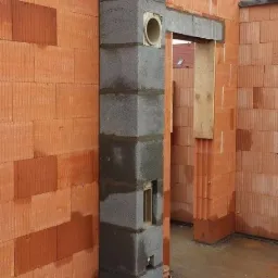 Budowa komina systemowego z szarych bloczków betonowych w surowym stanie, widoczny otwór wentylacyjny i fragmenty ścian z cegły ceramicznej