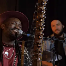 Muzyk w bordowym kapeluszu śpiewa do mikrofonu, grając na korze; w tle drugi muzyk przy perkusji.