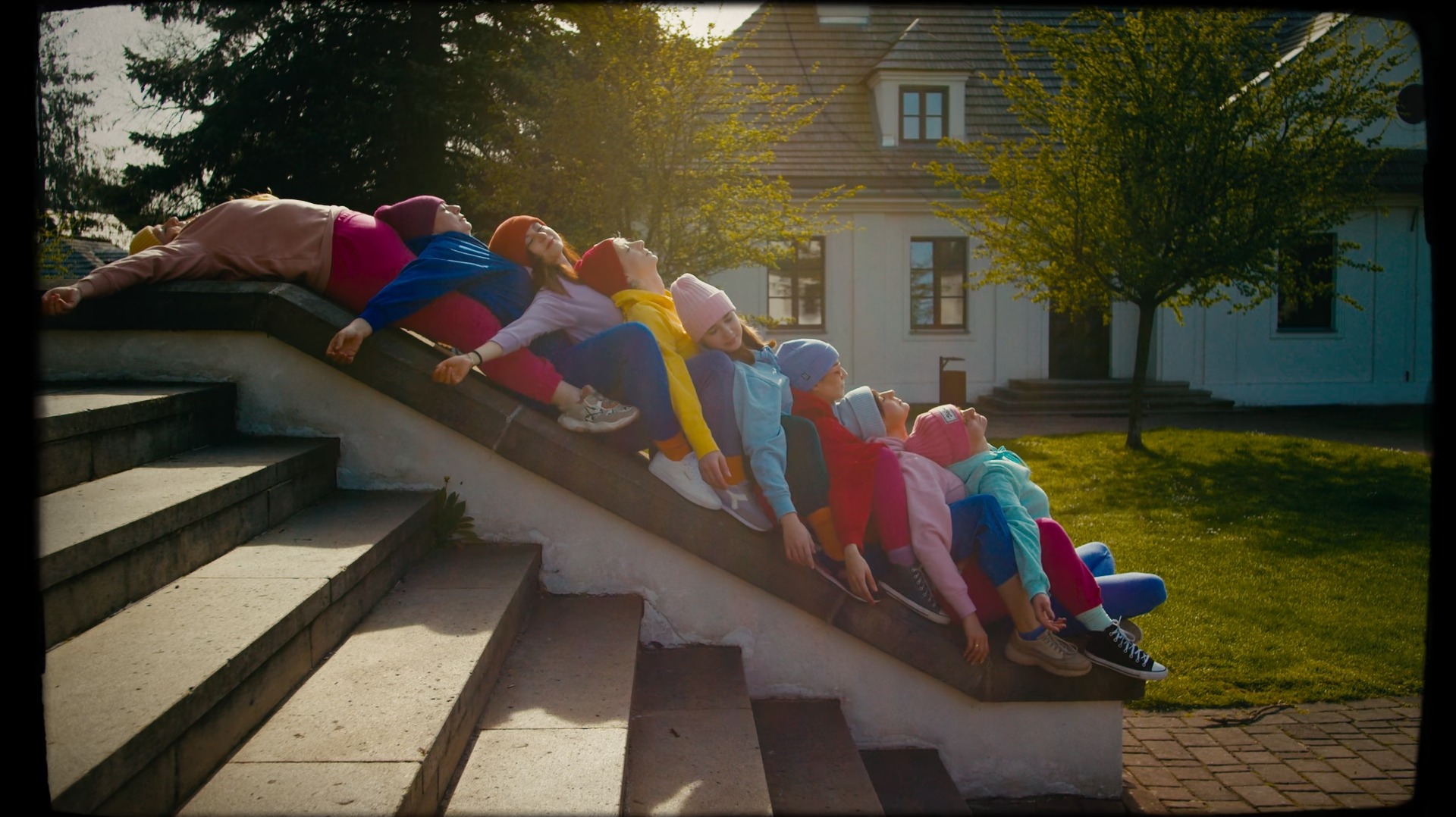 Kadr z planu filmowego: grupa młodych osób w barwnych ubraniach, leżących jeden za drugim na betonowych schodach, z budynkiem w tle, oświetlonych promieniami słońca.