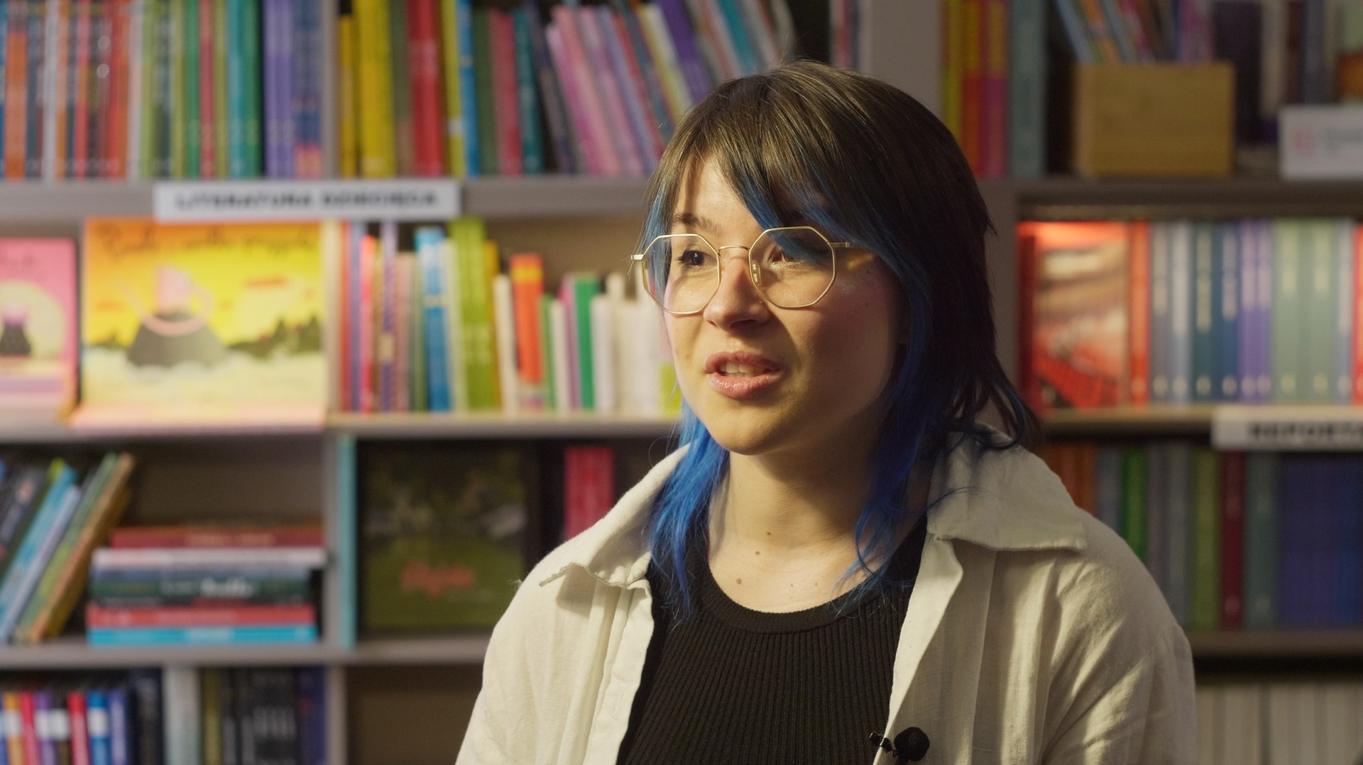 Kobieta w okularach z niebieskimi pasemkami w włosach mówi do kamery na tle półki z książkami.