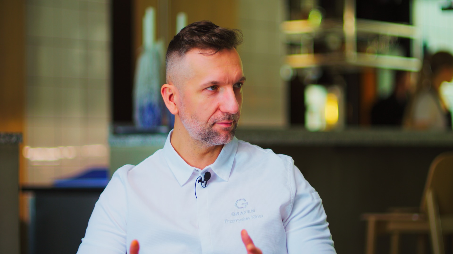 Mężczyzna w białej koszuli z mikrofonem krawatowym, logo firmy 'Grafen' widoczne na koszuli, gestykuluje dłońmi, w tle rozmyte wnętrze kawiarni.