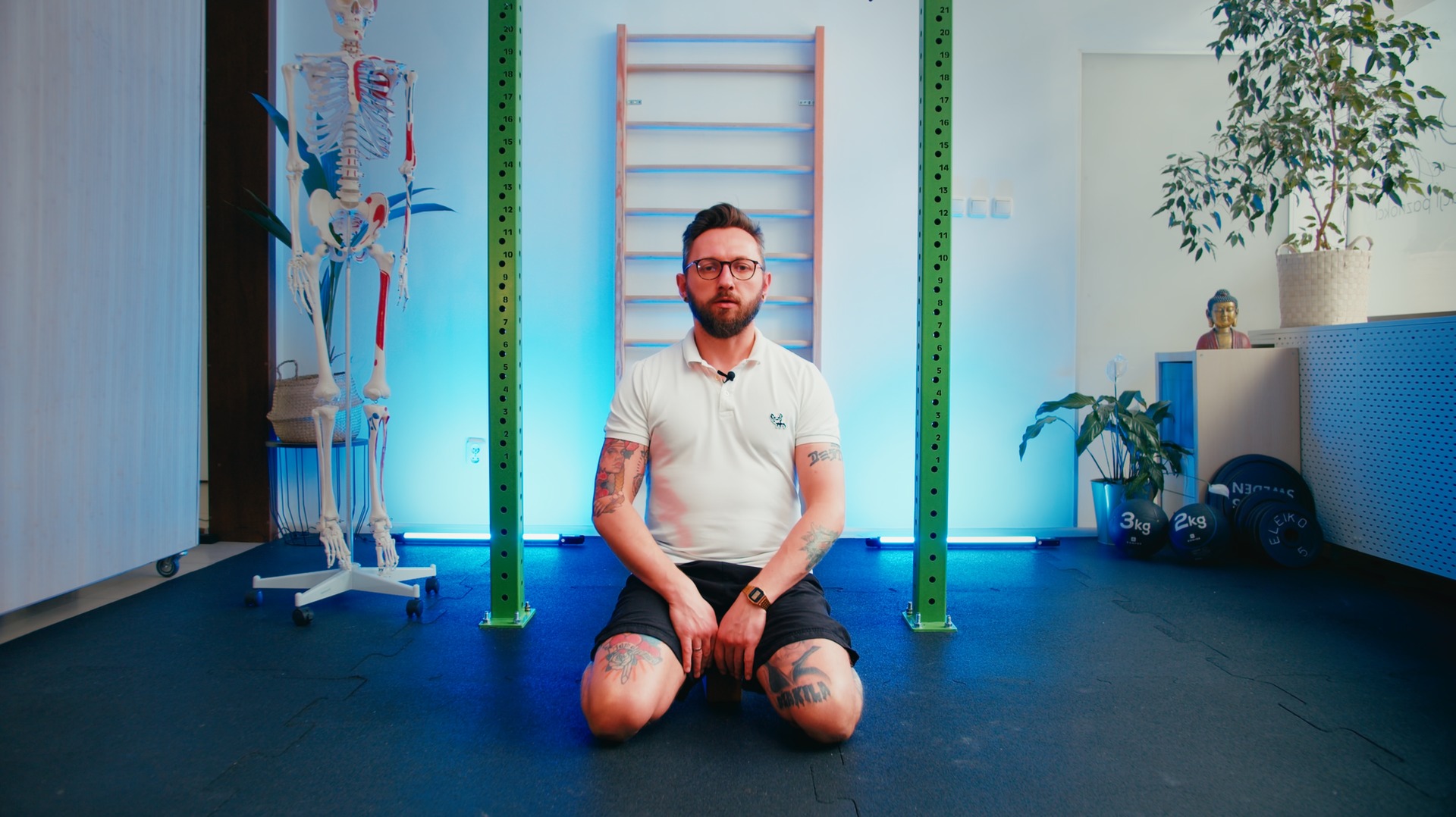 Mężczyzna w okularach klęczy w siłowni, prezentując ćwiczenia; widoczny szkielet anatomiczny, drabinki gimnastyczne i obciążenia.