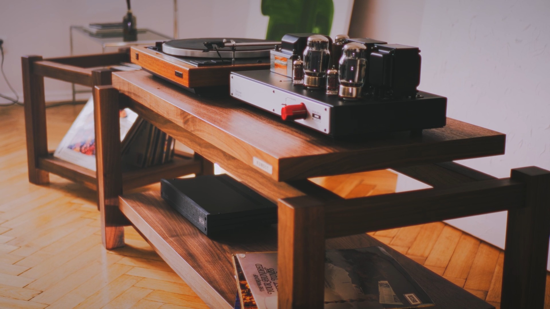 Drewniany stolik RTV z gramofonem, wzmacniaczem lampowym i płytami winylowymi, ustawiony na parkiecie w jodełkę.