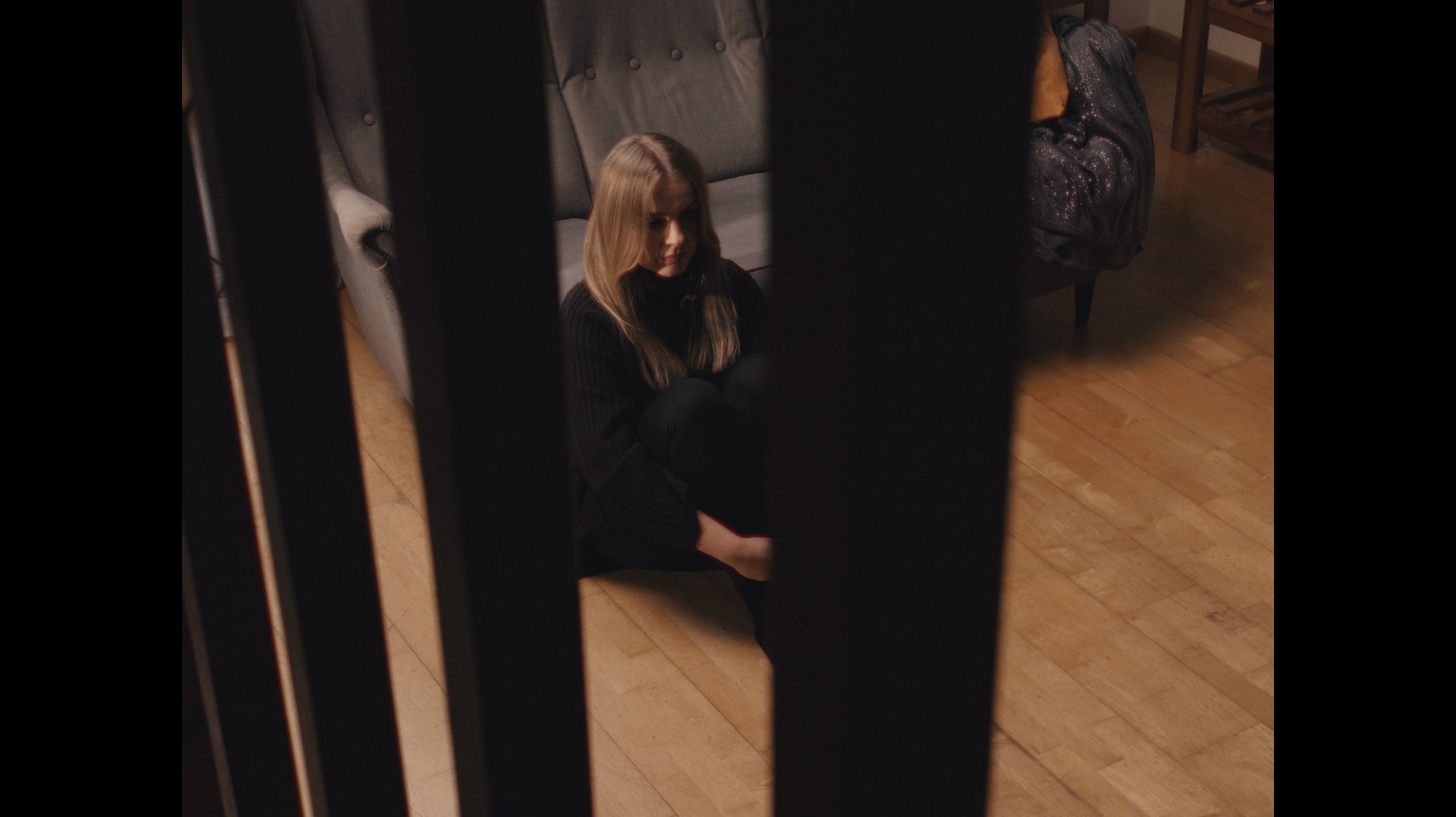 Młoda kobieta siedząca na podłodze, przytulona do kolan, widoczna przez ciemne, pionowe pręty; stonowana kolorystyka i melancholijny nastrój.