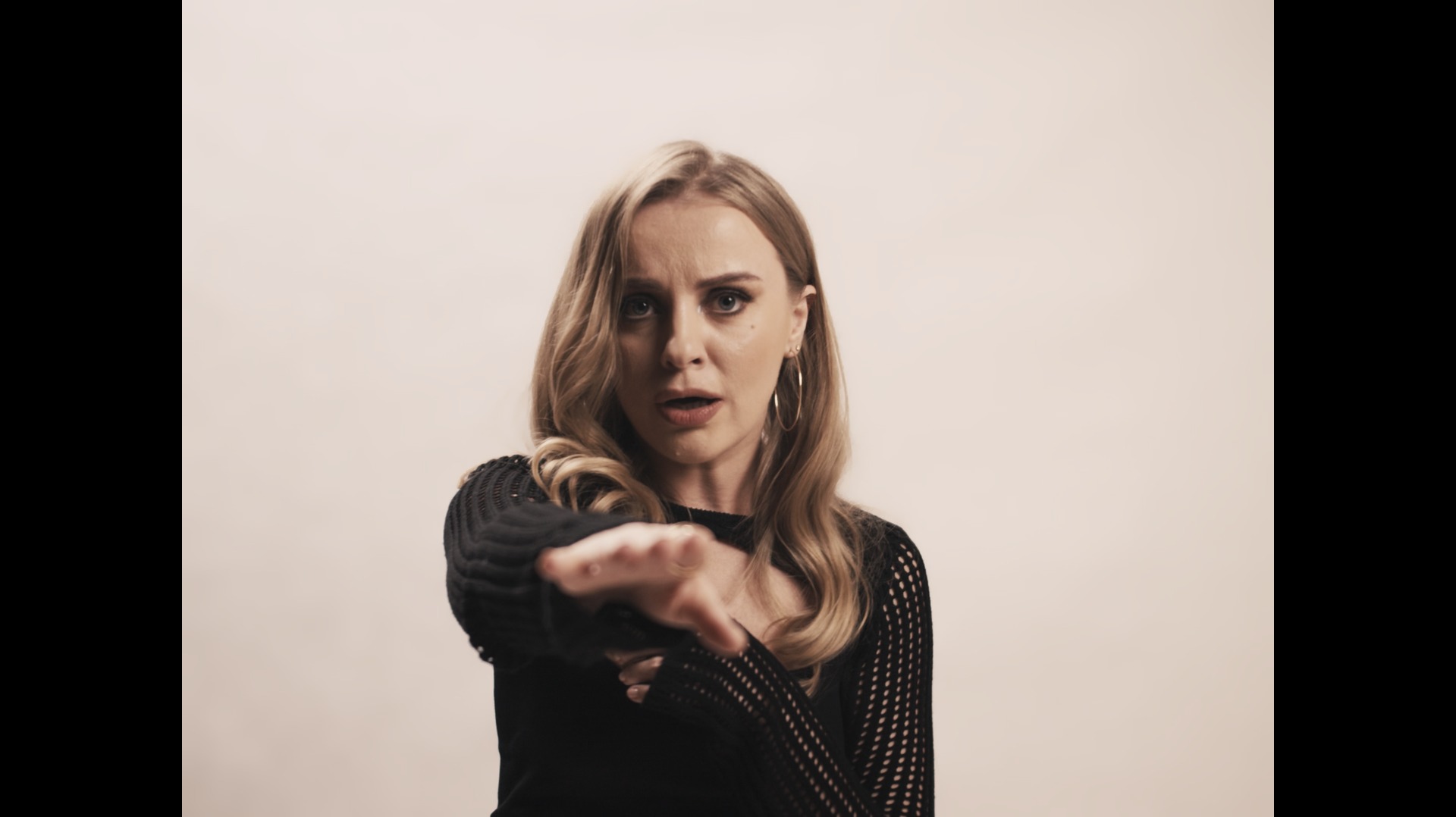 Blondwłosa kobieta w czarnej, ażurowej bluzce wyciąga rękę w kierunku kamery z zaskoczoną miną.