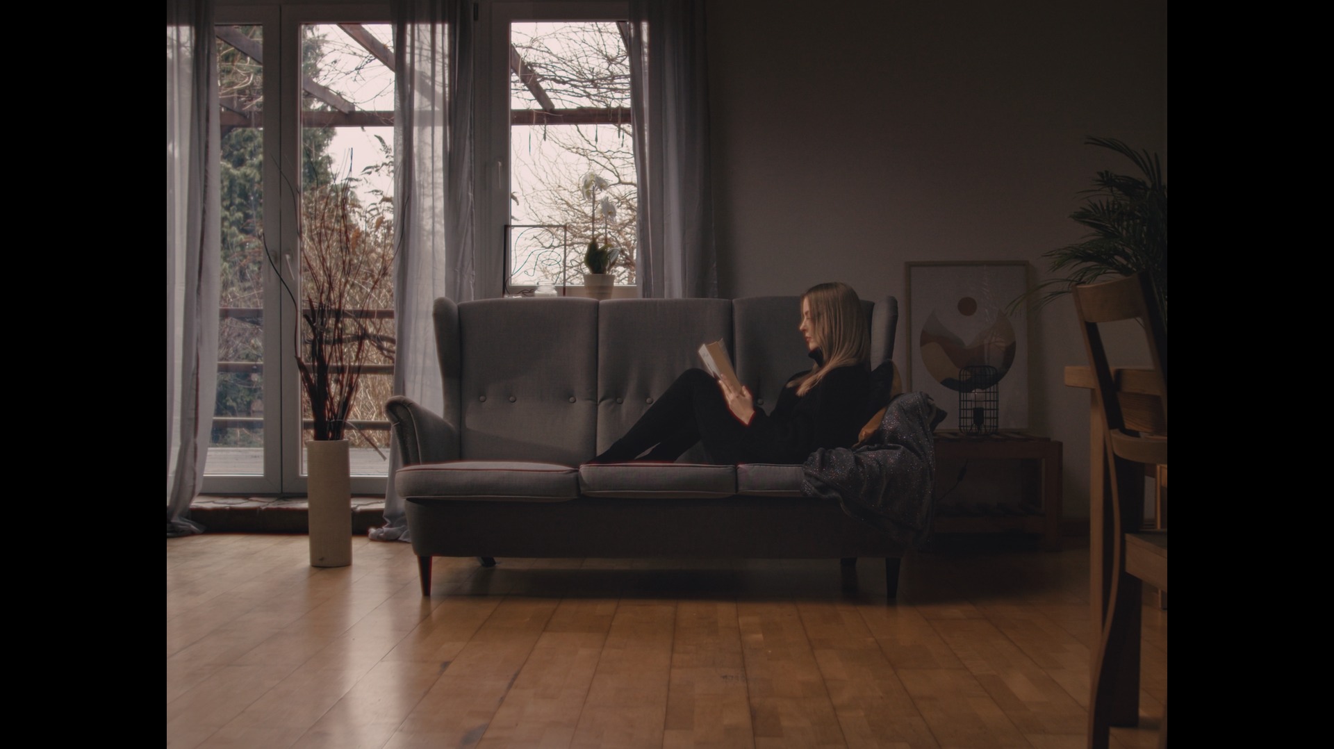 Kadr z filmu przedstawiający kobietę czytającą książkę na szarej sofie w salonie z dużym oknem i drewnianą podłogą.