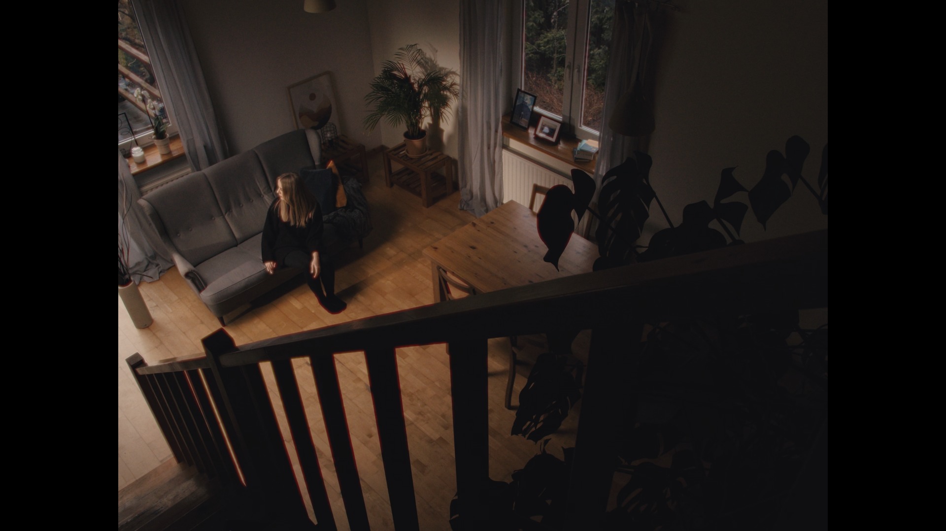 Kobieta siedząca na szarej kanapie w salonie w Krakowie, widok z góry schodów, z widocznymi roślinami doniczkowymi i drewnianym stołem.