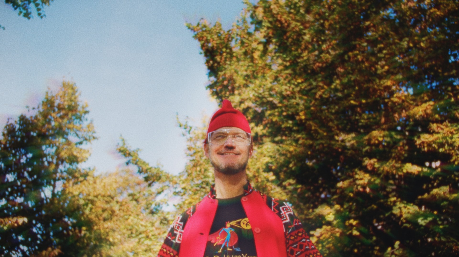Portret uśmiechniętego mężczyzny w czerwonej czapce skrzata i okularach ochronnych, ubrany w kolorową koszulkę z nadrukiem superbohatera i czerwoną kamizelkę, na tle drzew i błękitnego nieba.