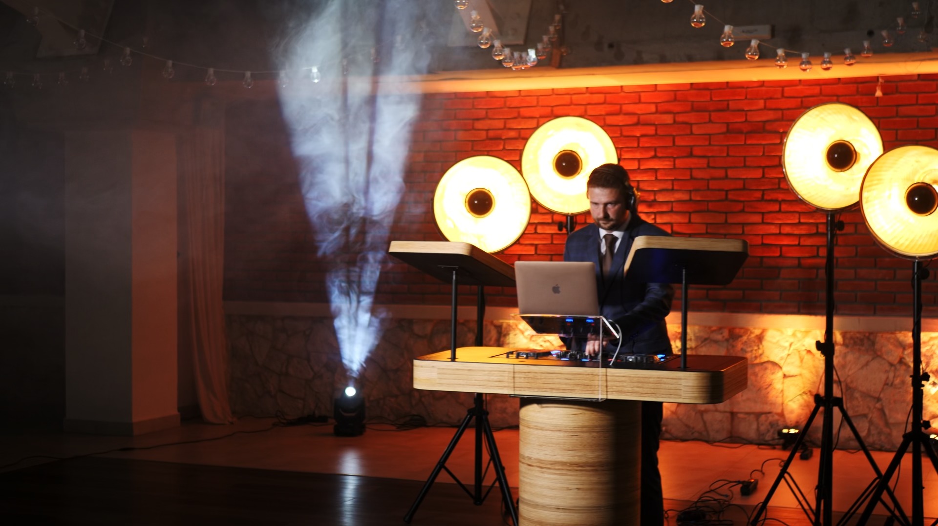 DJ w garniturze obsługujący sprzęt podczas imprezy, oświetlony reflektorami i z efektem dymu, na tle ściany z czerwonej cegły.