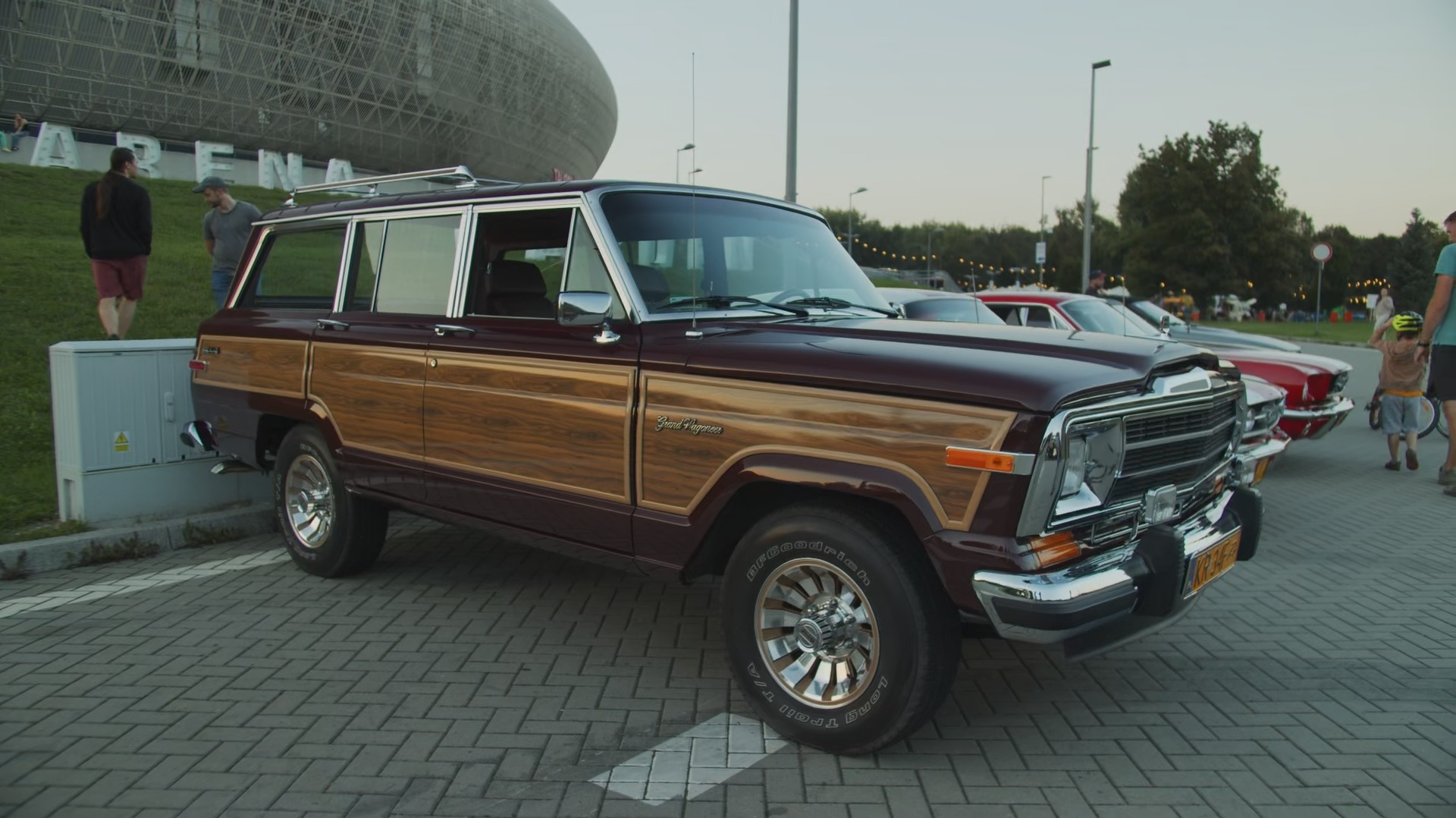Burgundowy Jeep Grand Wagoneer z drewnianymi panelami bocznymi, zaparkowany na brukowanym placu, z widocznymi innymi klasycznymi autami w tle i fragmentem nowoczesnej architektury.