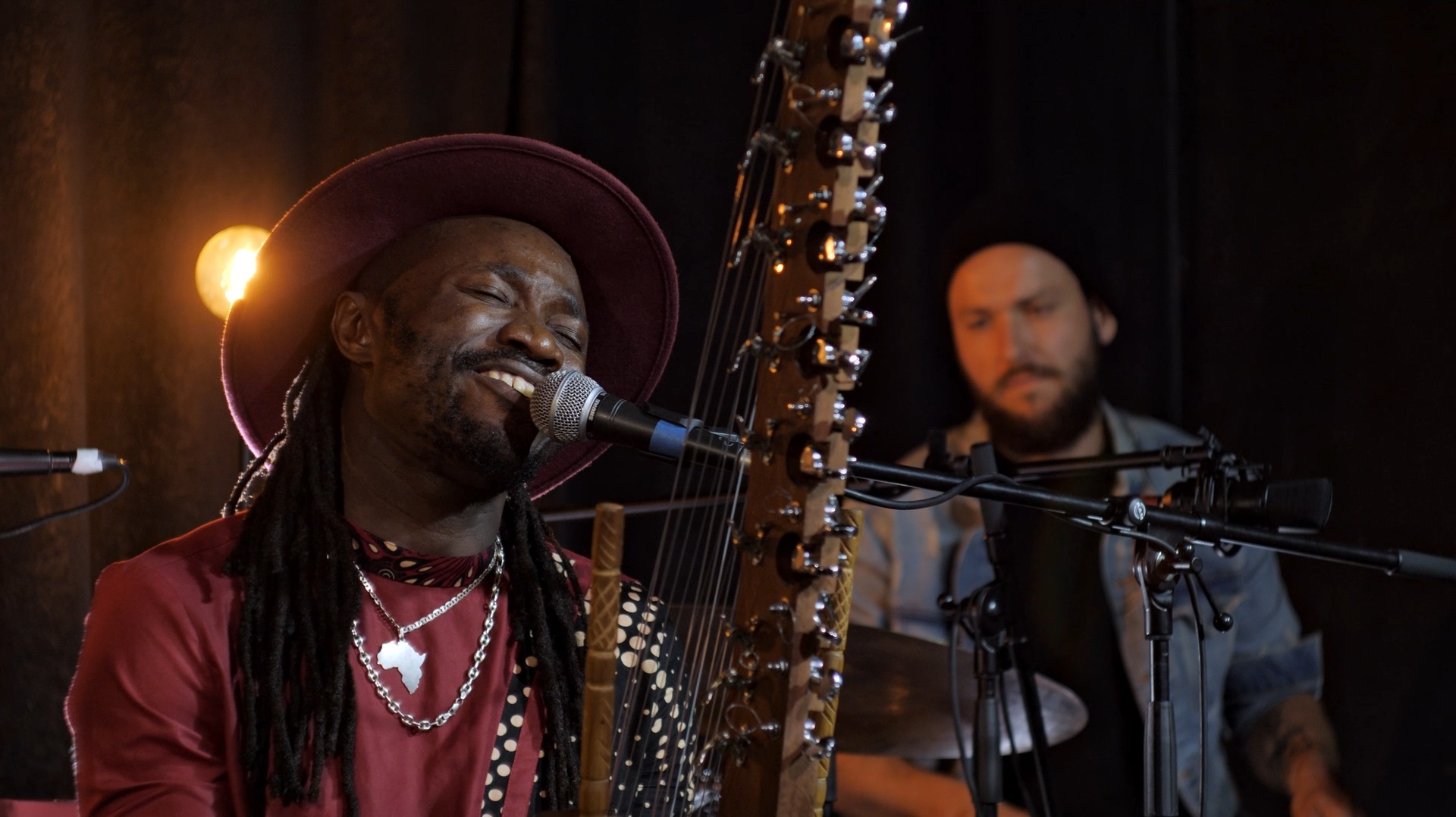 Muzyk w bordowym kapeluszu śpiewa do mikrofonu, grając na korze; w tle drugi muzyk przy perkusji.