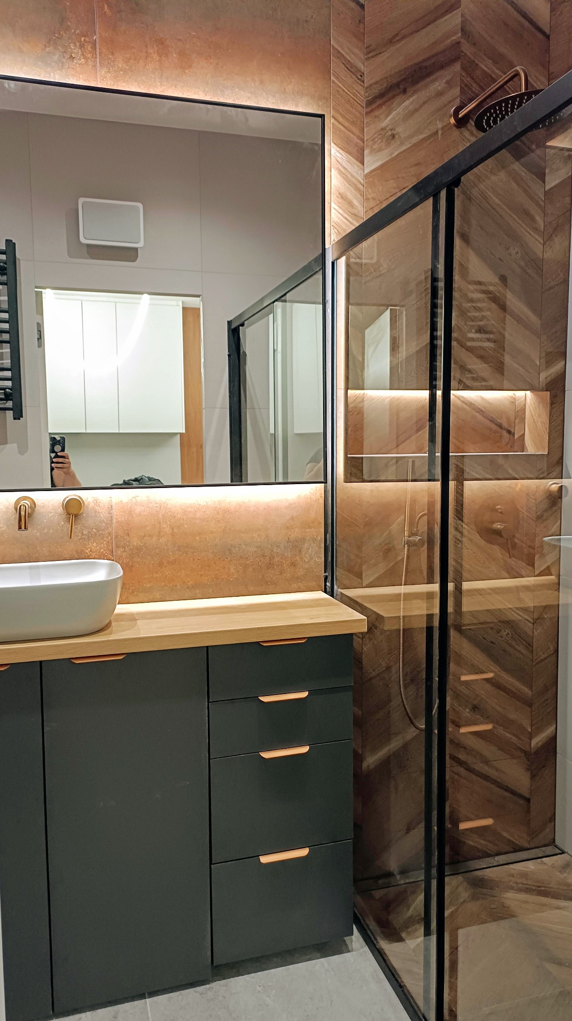 Nowoczesna łazienka: szara szafka z drewnianym blatem, owalna umywalka, lustro z podświetleniem i kabina prysznicowa z drewnopodobną okładziną.