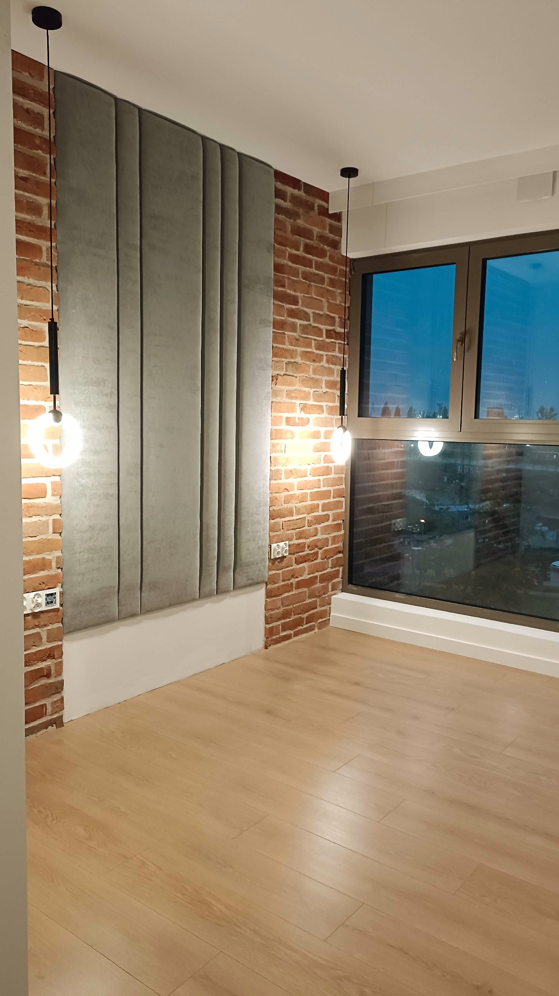 Pokój z ceglaną ścianą i panelami ściennymi, oświetlony wiszącymi lampami. Widoczne gniazdka elektryczne i drewniana podłoga.