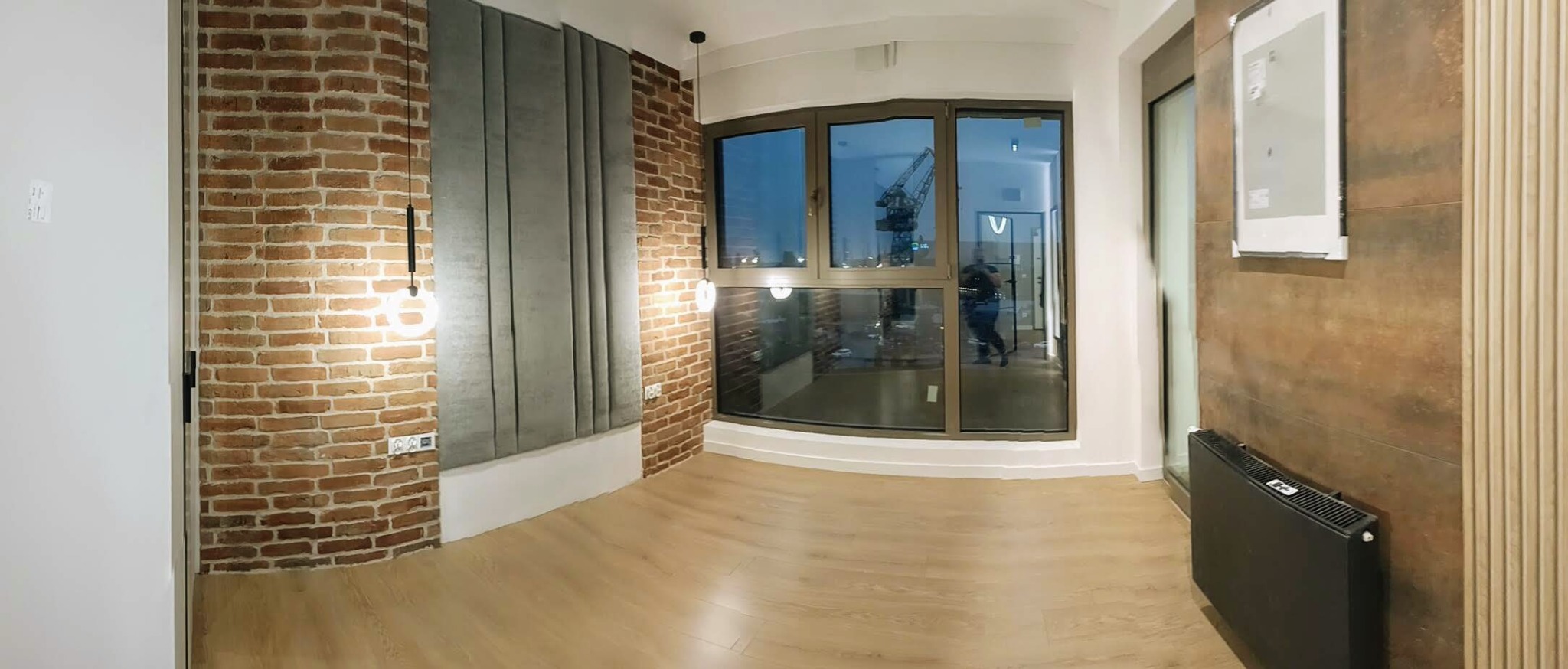 Nowoczesne wykończenie wnętrza: ceglana ściana, szare panele tapicerowane, drewniana podłoga i okno z widokiem na miasto. Elementy oświetlenia.