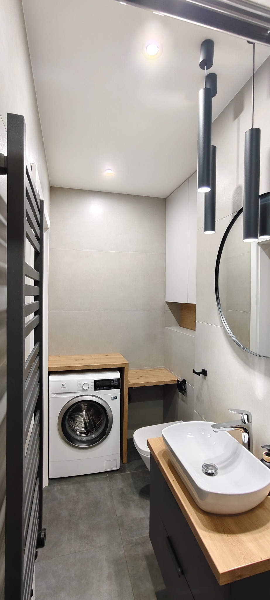 Nowoczesna łazienka z pralką zabudowaną w drewnianej obudowie, okrągłym lustrem i podwieszanymi lampami cylindrycznymi. Minimalistyczny design, szaro-biała kolorystyka.
