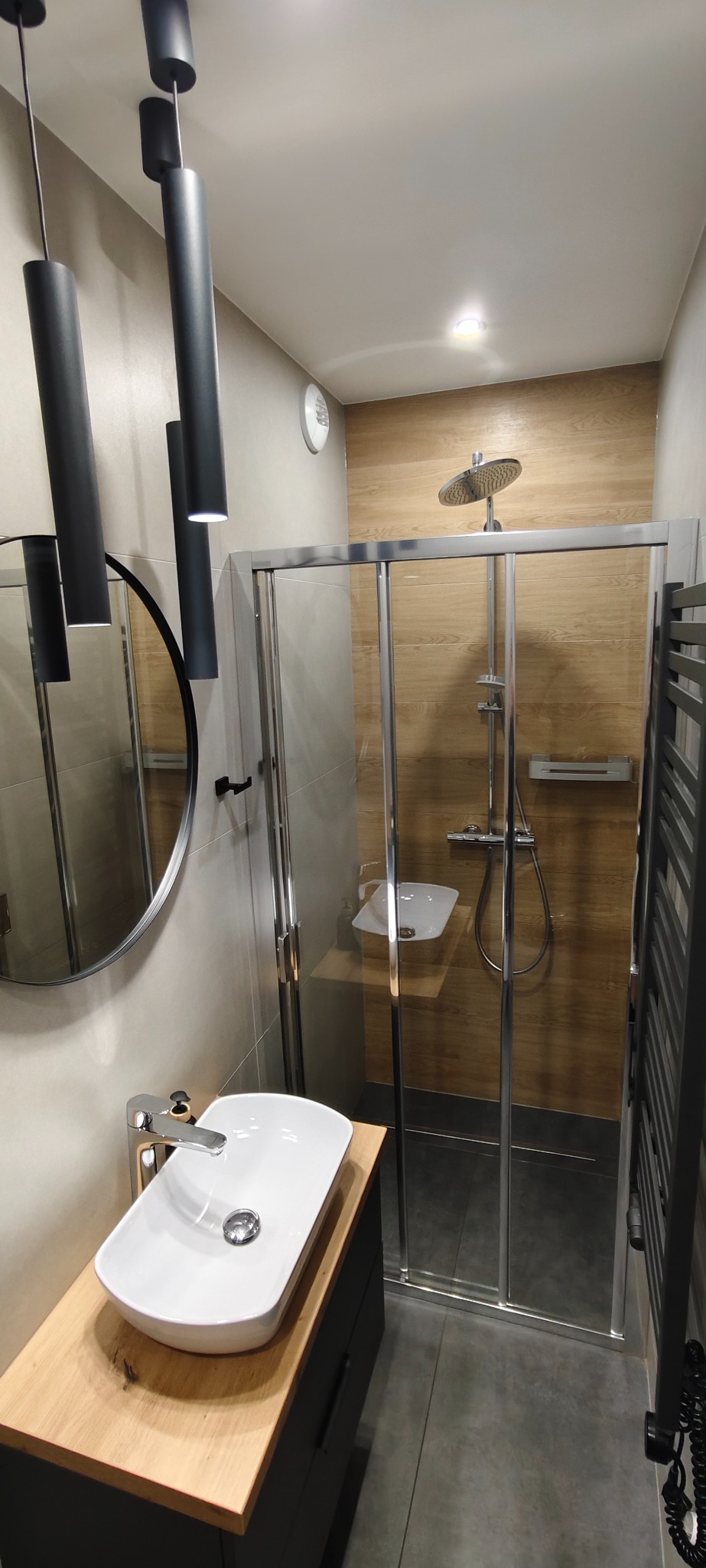 Nowoczesna łazienka z kabiną prysznicową, umywalką nablatową i okrągłym lustrem. Minimalistyczne oświetlenie, drewniane akcenty i szara kolorystyka.