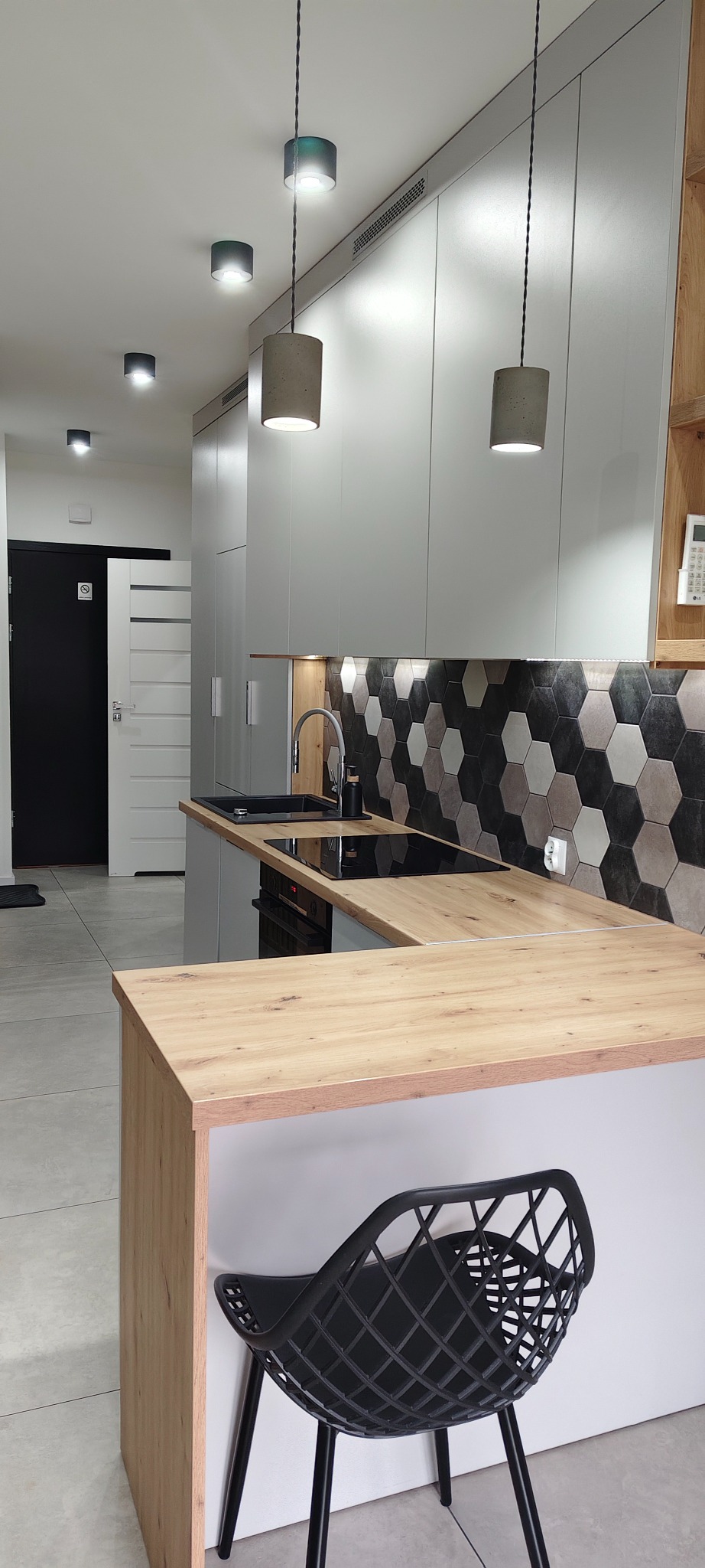 Nowoczesna kuchnia z heksagonalnymi płytkami, drewnianym blatem i designerskimi lampami. Minimalistyczne szafki w szarościach, czarne krzesło barowe.