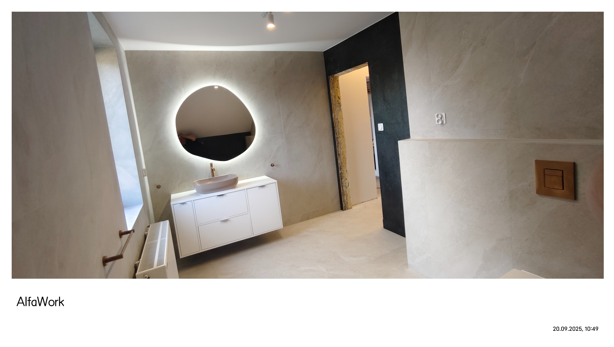 Elegancka łazienka z podświetlanym lustrem o nieregularnym kształcie, umywalką w kolorze beżu i białą szafką. Na ścianach płytki imitujące kamień, widoczny grzejnik i fragment drzwi.