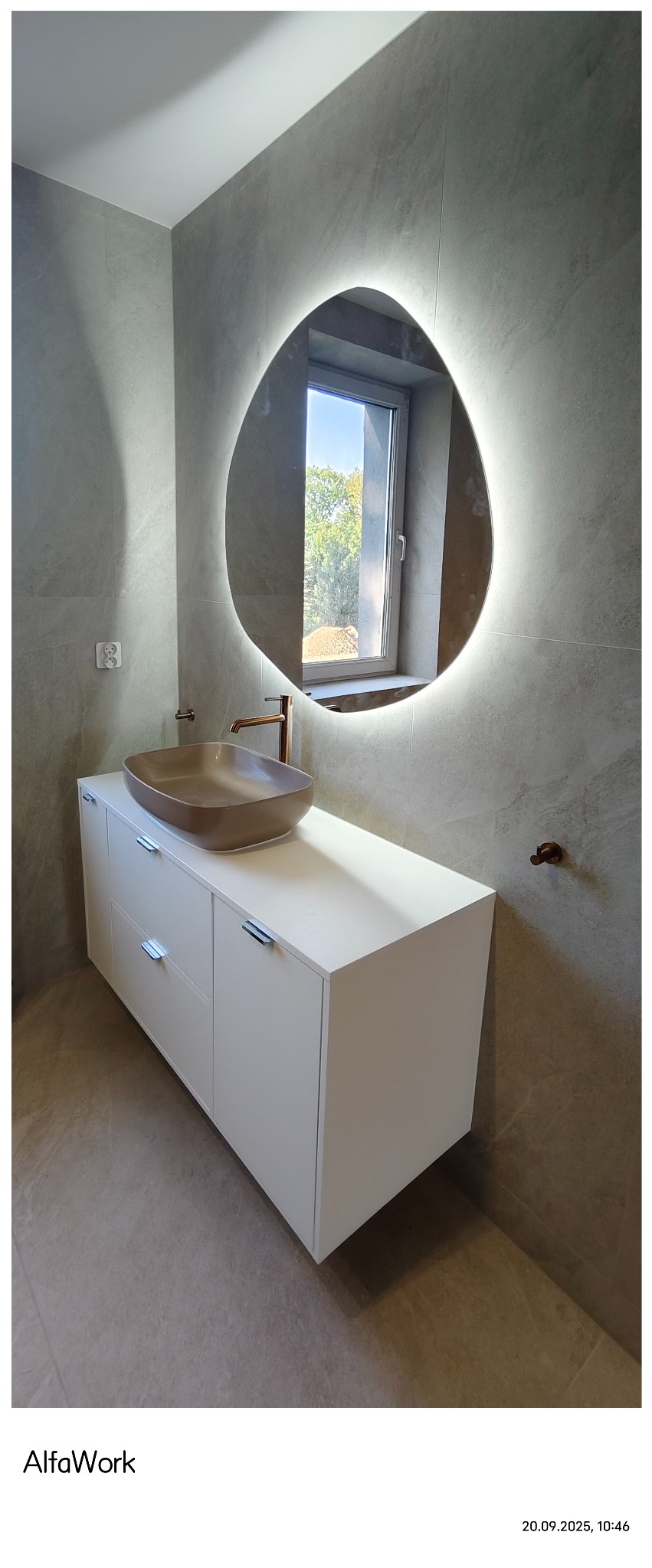 Nowoczesna łazienka z umywalką w kolorze taupe i podświetlanym, asymetrycznym lustrem. Minimalistyczna szafka pod umywalką i szare płytki na ścianach.