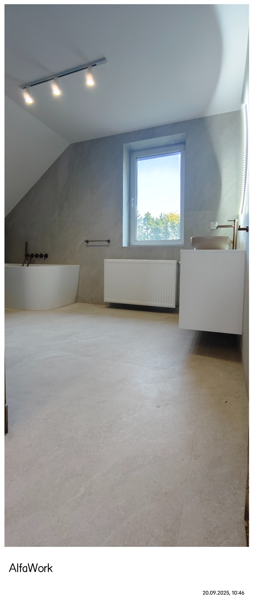 Jasna łazienka na poddaszu z beżowymi płytkami, wanną, grzejnikiem i umywalką. Okno z widokiem na drzewa. Oświetlenie punktowe.