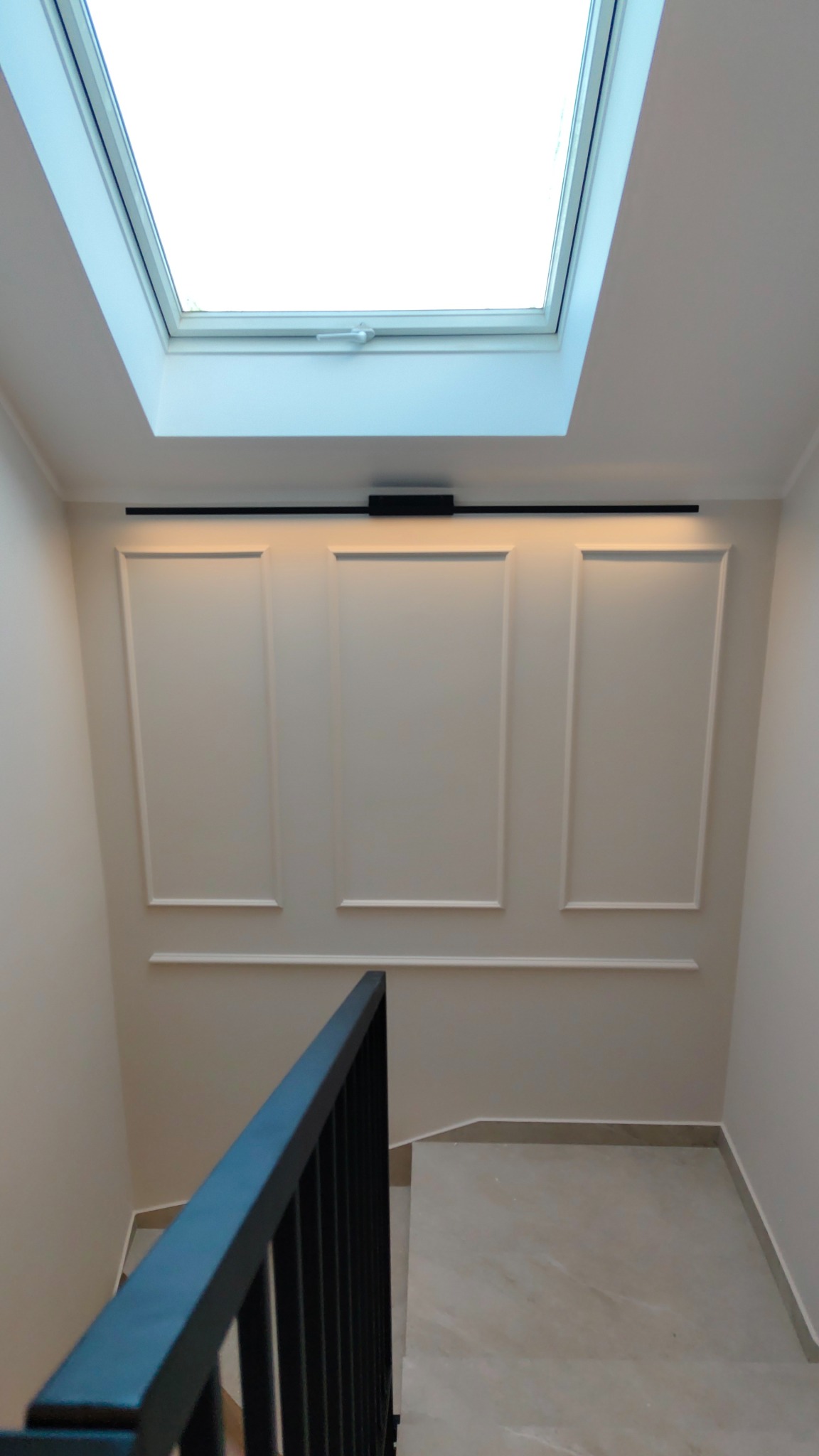 Klatka schodowa z beżowymi ścianami, dekoracyjnymi panelami, czarnym oświetleniem i balustradą. W suficie okno dachowe wpuszczające naturalne światło.