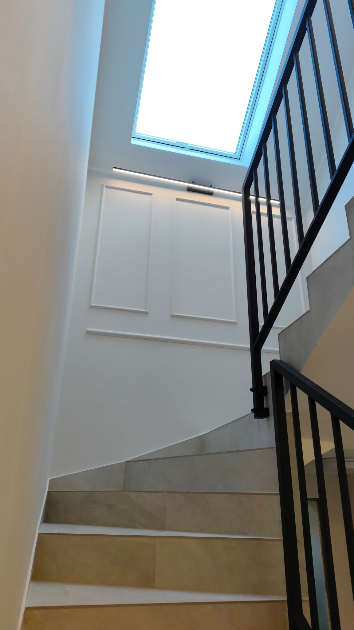 Białe ściany z dekoracyjnymi panelami i oświetleniem LED, schody wyłożone płytkami oraz czarna balustrada podświetlone przez okno dachowe.