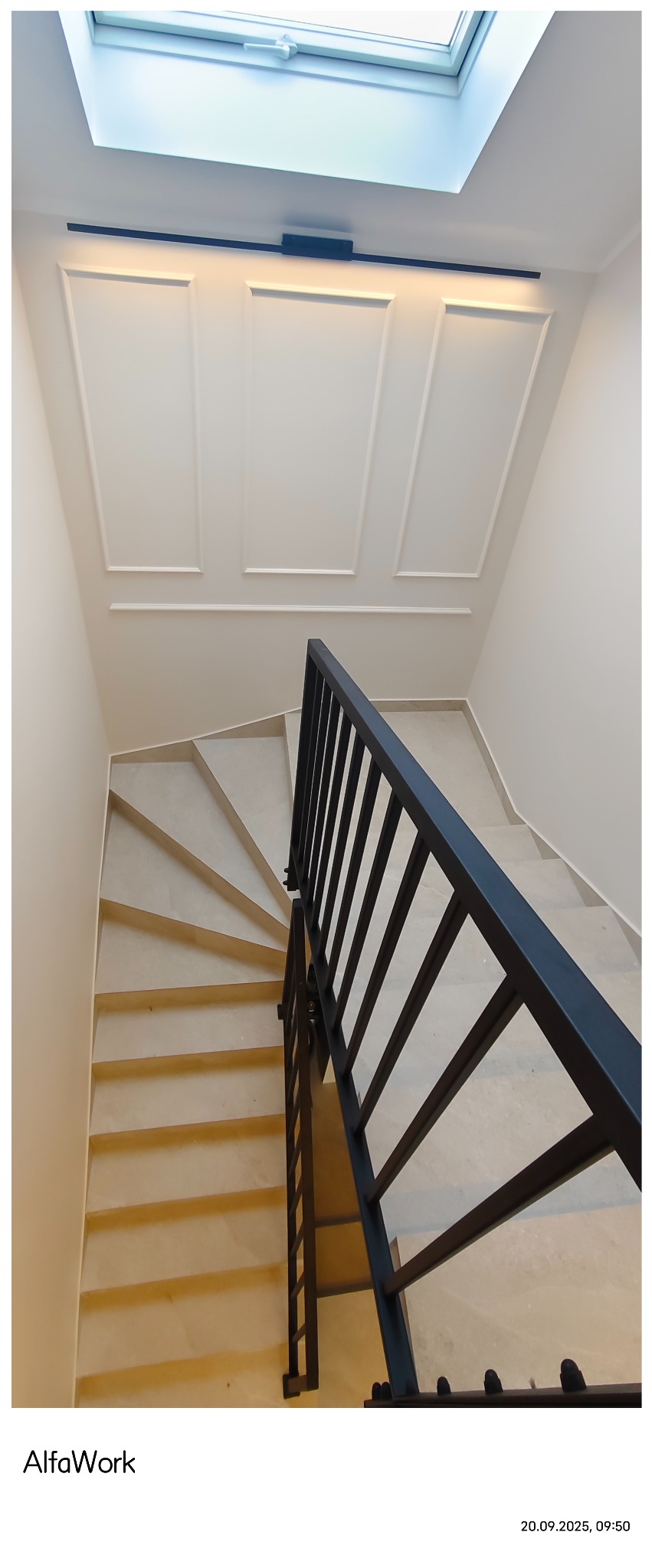 Klatka schodowa wykończona w jasnych barwach z czarną balustradą. Widok z góry na spiralne schody z kamiennymi stopniami. Na ścianie dekoracyjne panele.