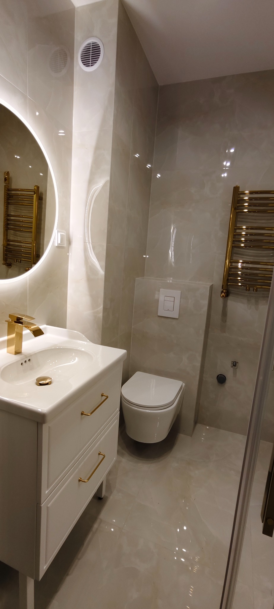 Elegancka łazienka w beżowych płytkach z białą umywalką, złotą armaturą i grzejnikiem. Okrągłe lustro z podświetleniem LED. Minimalistyczna toaleta podwieszana.