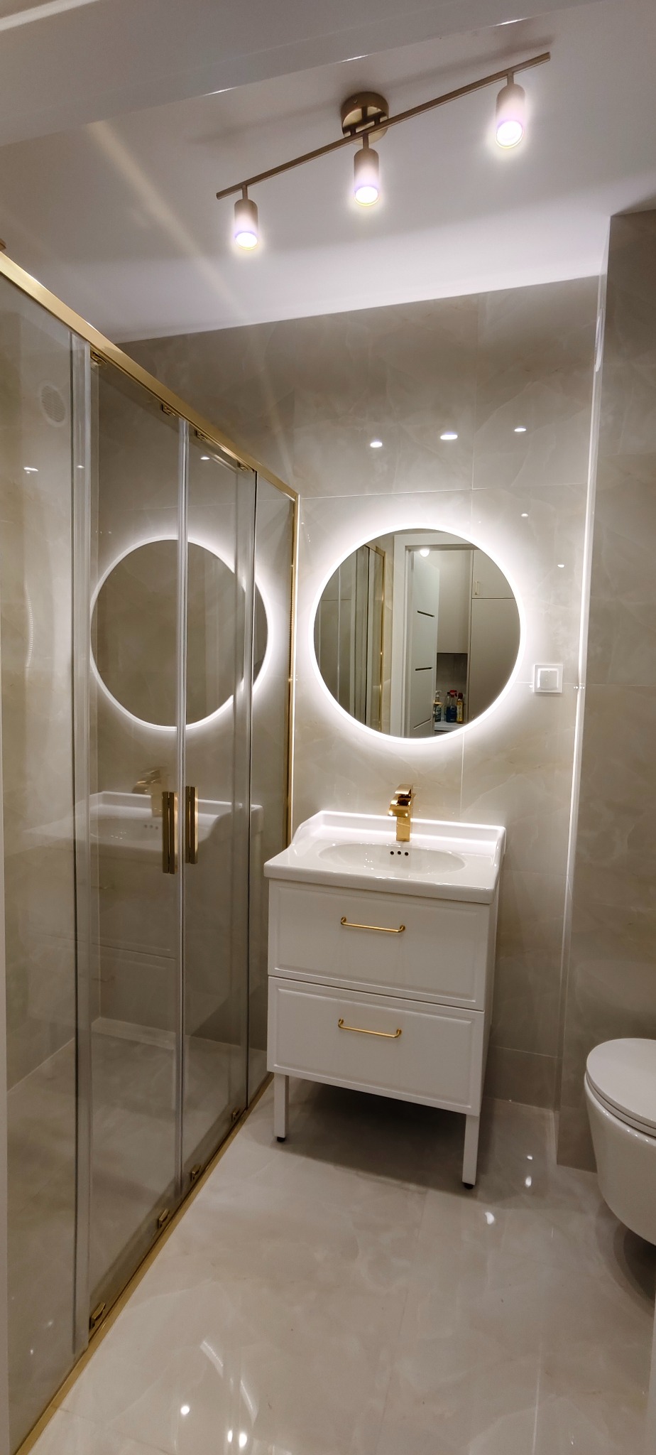 Wykończona łazienka z kabiną prysznicową ze złotymi ramami, białą szafką z umywalką i okrągłym lustrem z podświetleniem LED. Beżowe płytki na ścianach i podłodze.