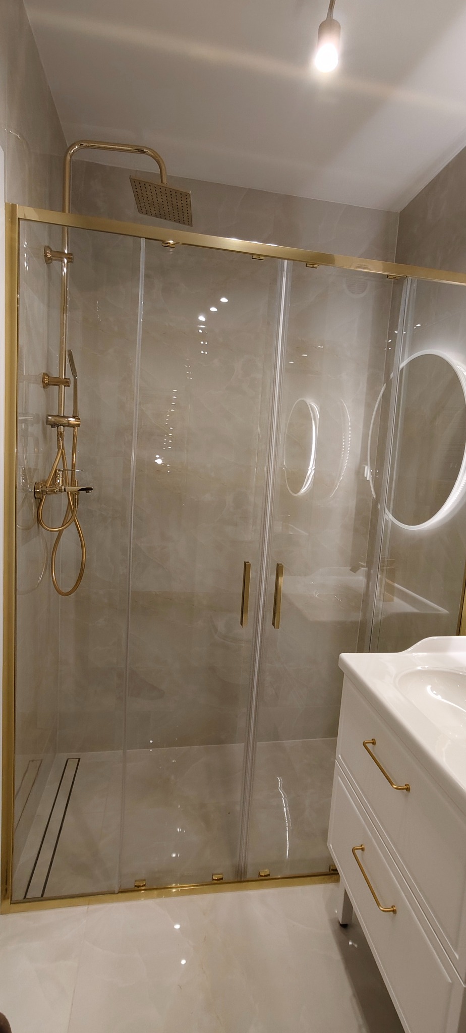 Nowoczesna kabina prysznicowa z złotymi detalami, widoczna umywalka z szafką w jasnej łazience. Minimalistyczny design z okrągłym lustrem z podświetleniem.