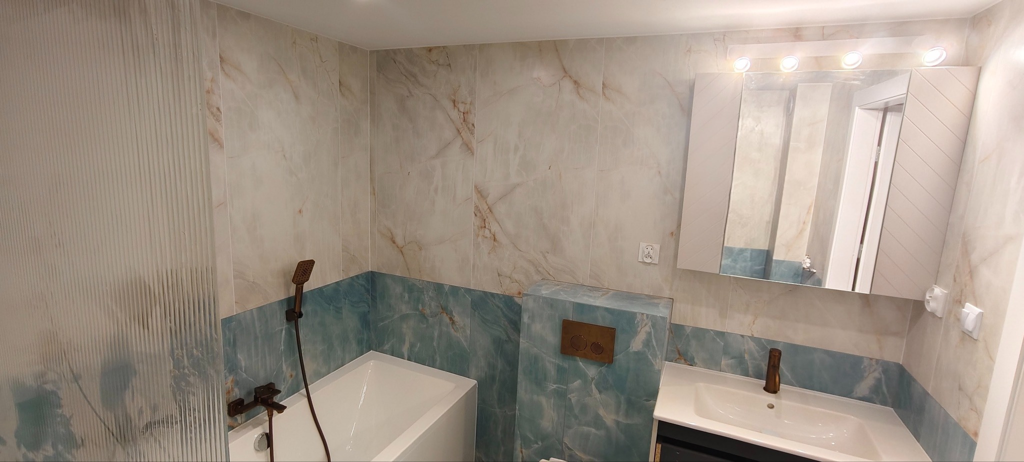 Wykończona łazienka z wanną, umywalką i toaletą w zabudowie. Płytki imitujące marmur w odcieniach beżu i błękitu. Armatura w kolorze starego złota. Nowoczesny design.