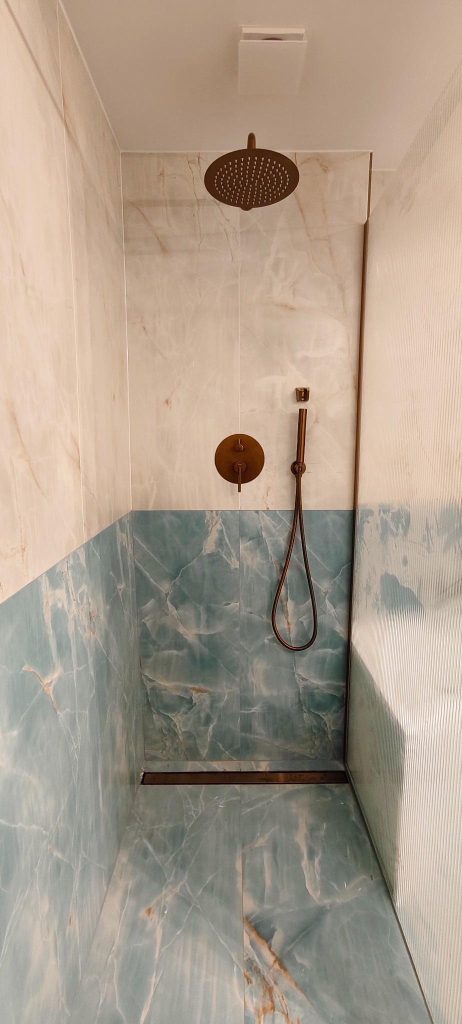 Wykończona kabina prysznicowa z marmurowymi płytkami w odcieniach bieli i błękitu, z deszczownicą i baterią w kolorze mosiądzu. Nowoczesny design z elementami retro.