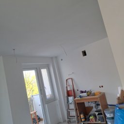 Kewbud - Pokój w trakcie odświeżania: świeżo pomalowane ściany na biało, widoczne narzędzia malarskie, drabina i prowizoryczny stół roboczy z farbami, przygotowany do dalszych prac wykończeniowych.