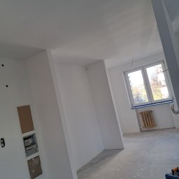 Kewbud - Wnętrze w trakcie prac wykończeniowych we Wrocławiu: świeżo pomalowane na biało ściany, widoczne puszki elektryczne, grzejnik pod oknem i rozłożone przewody na podłodze.