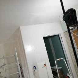 Kewbud - Wnętrze w trakcie prac wykończeniowych: ściany pokryte świeżo nałożoną gładzią, widoczne puszki elektryczne, drabiny i sprzęt oświetleniowy, przygotowane do dalszego malowania.