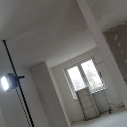 Kewbud - Wnętrze pokoju w trakcie prac wykończeniowych, widoczna lampa budowlana na statywie, okno z widokiem na zewnątrz, ściany i sufit w trakcie przygotowania do malowania, częściowo pokryte gładzią,...