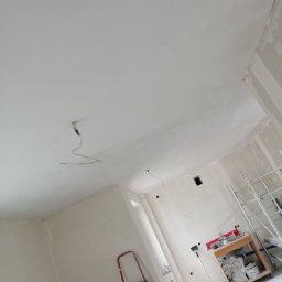 Kewbud - Surowe wnętrze w trakcie szpachlowania ścian i sufitu, widoczne narzędzia, drabina malarska i przygotowane materiały budowlane.