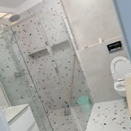 Nowoczesna łazienka z kabiną prysznicową typu walk-in, umywalką z szafką i toaletą w zabudowie, wykończona płytkami w stylu terrazzo oraz gładkimi, jasnymi płytkami na ścianach.