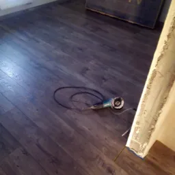 Układanie paneli podłogowych z ciemnego drewna, widoczna szlifierka kątowa przy ścianie i karton po telewizorze UHD TV w tle.