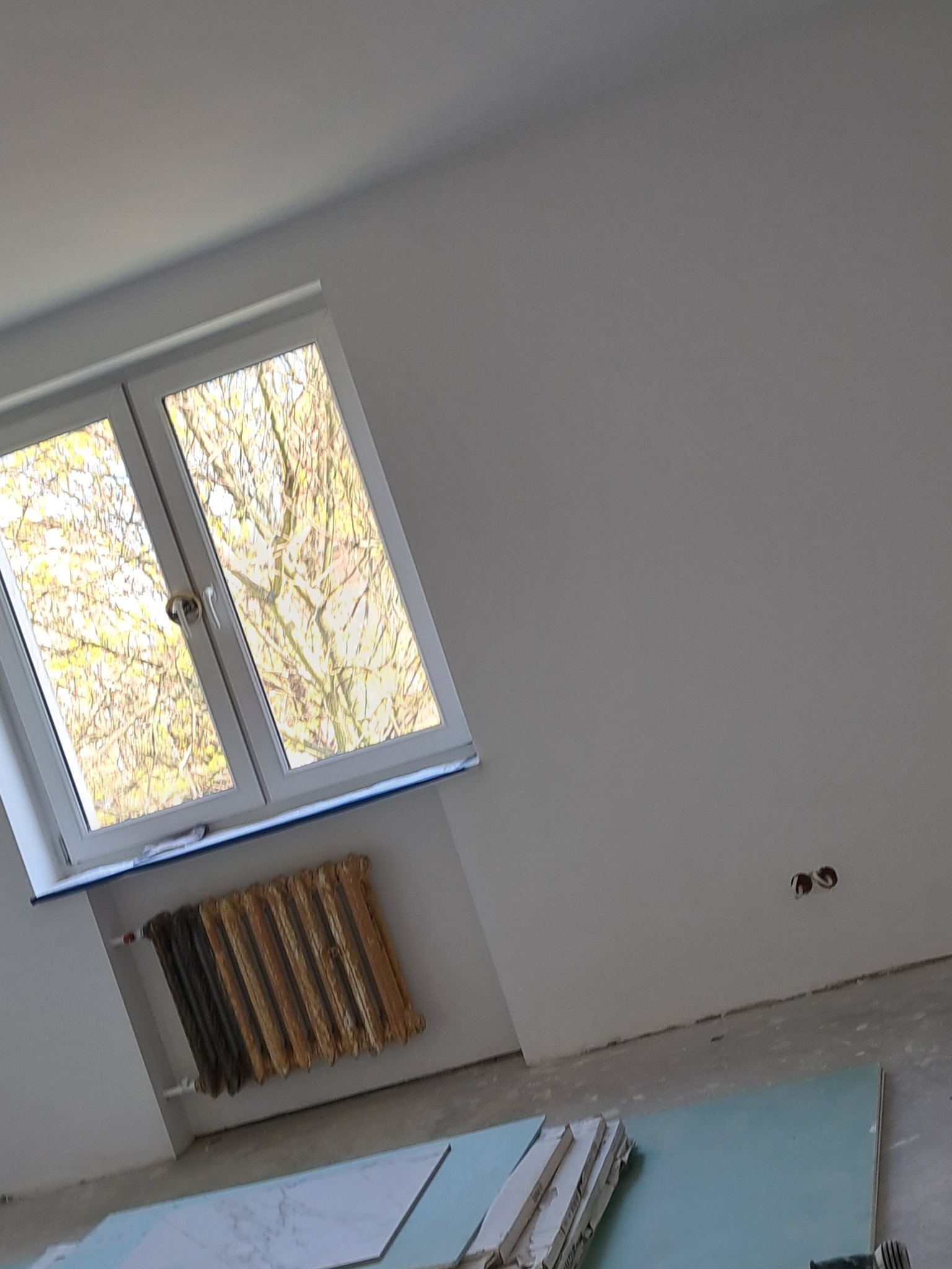 Wnętrze w trakcie remontu: widok na okno z widokiem na drzewo, stary, częściowo pomalowany grzejnik, gładka ściana z gniazdami elektrycznymi, płyty gipsowo-kartonowe i płytki podłogowe.