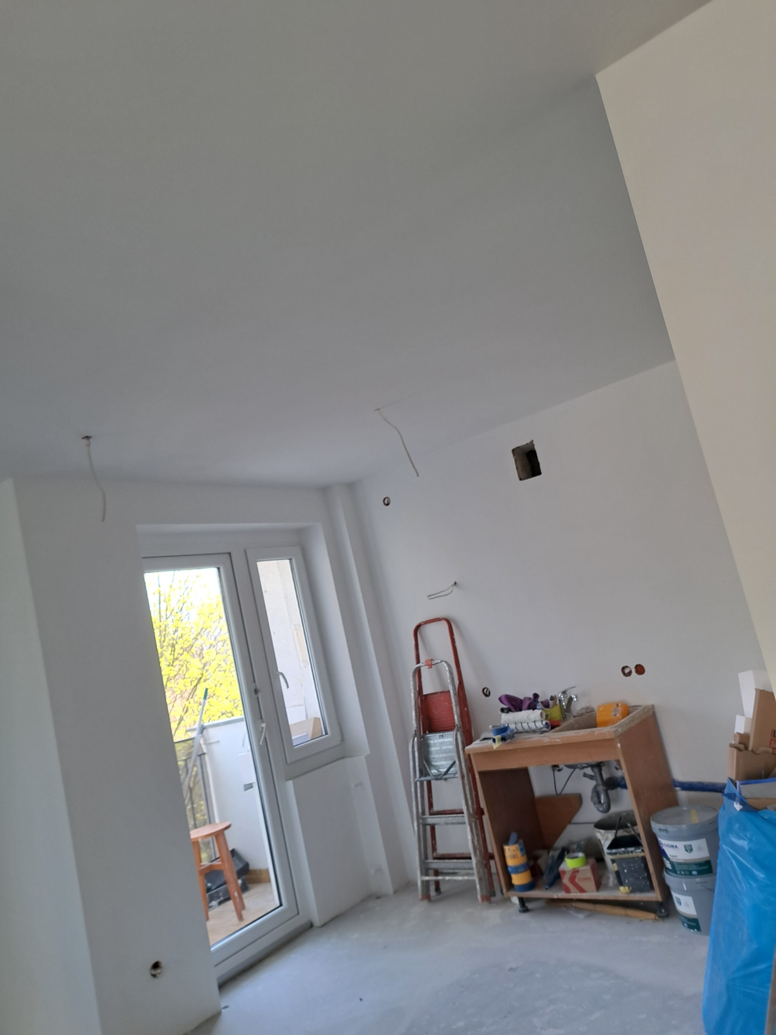 Pokój w trakcie odświeżania: świeżo pomalowane ściany na biało, widoczne narzędzia malarskie, drabina i prowizoryczny stół roboczy z farbami, przygotowany do dalszych prac wykończeniowych.