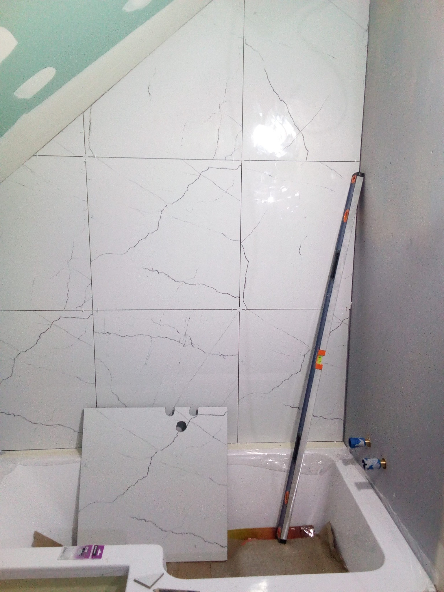 Białe płytki imitujące marmur na ścianie nad wanną, częściowo ułożone, z poziomnicą opartą o ścianę i niezamontowaną płytką z otworami na baterię.