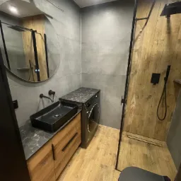 Nowoczesna łazienka z kabiną prysznicową typu walk-in, umywalką nablatową i pralką pod blatem. Minimalistyczny design z drewnem i szarością.