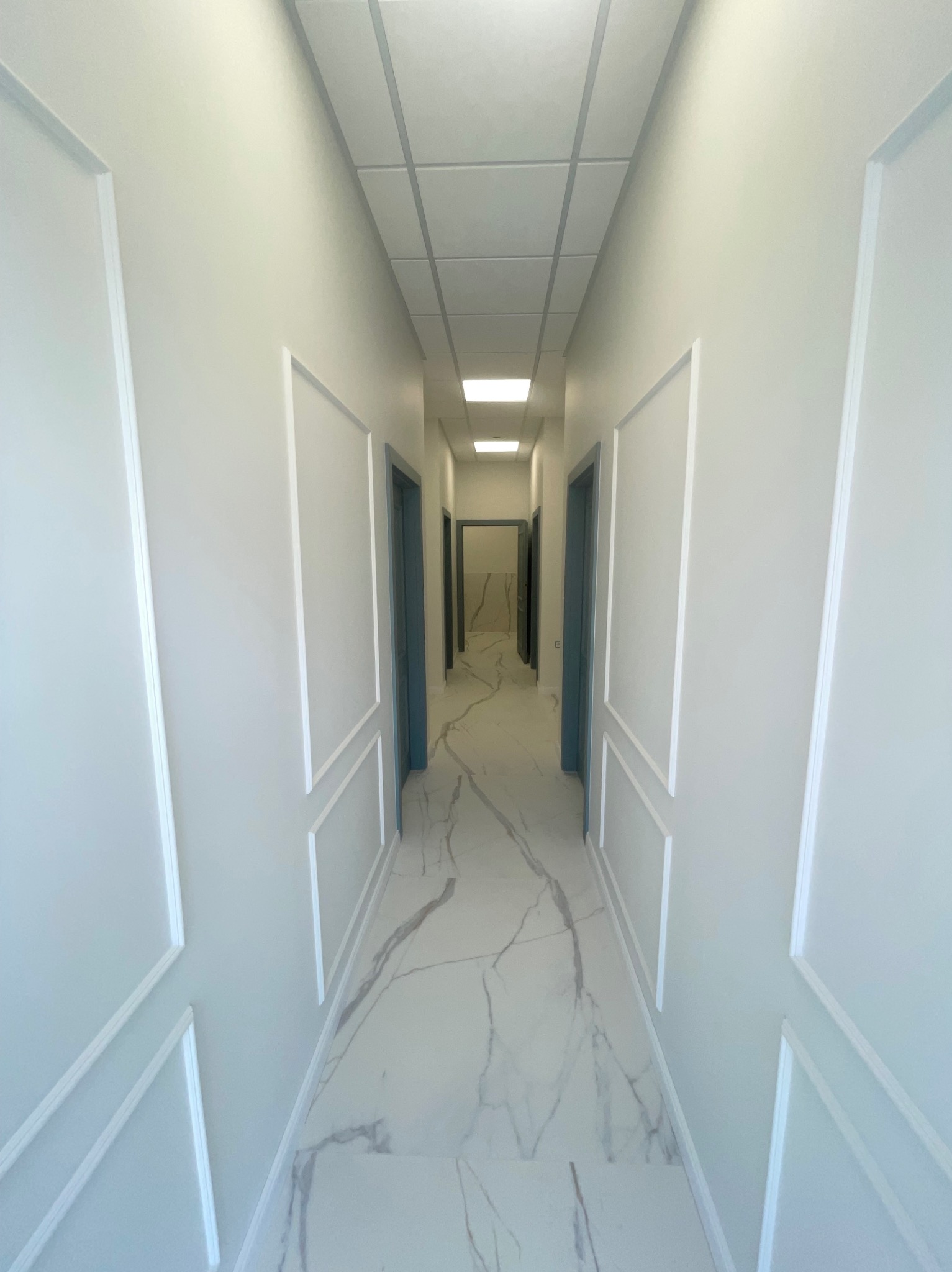 Korytarz w jasnych barwach z marmurową podłogą i dekoracyjnymi panelami ściennymi. Widoczne drzwi w kolorze szaro-niebieskim i sufit kasetonowy z oświetleniem.
