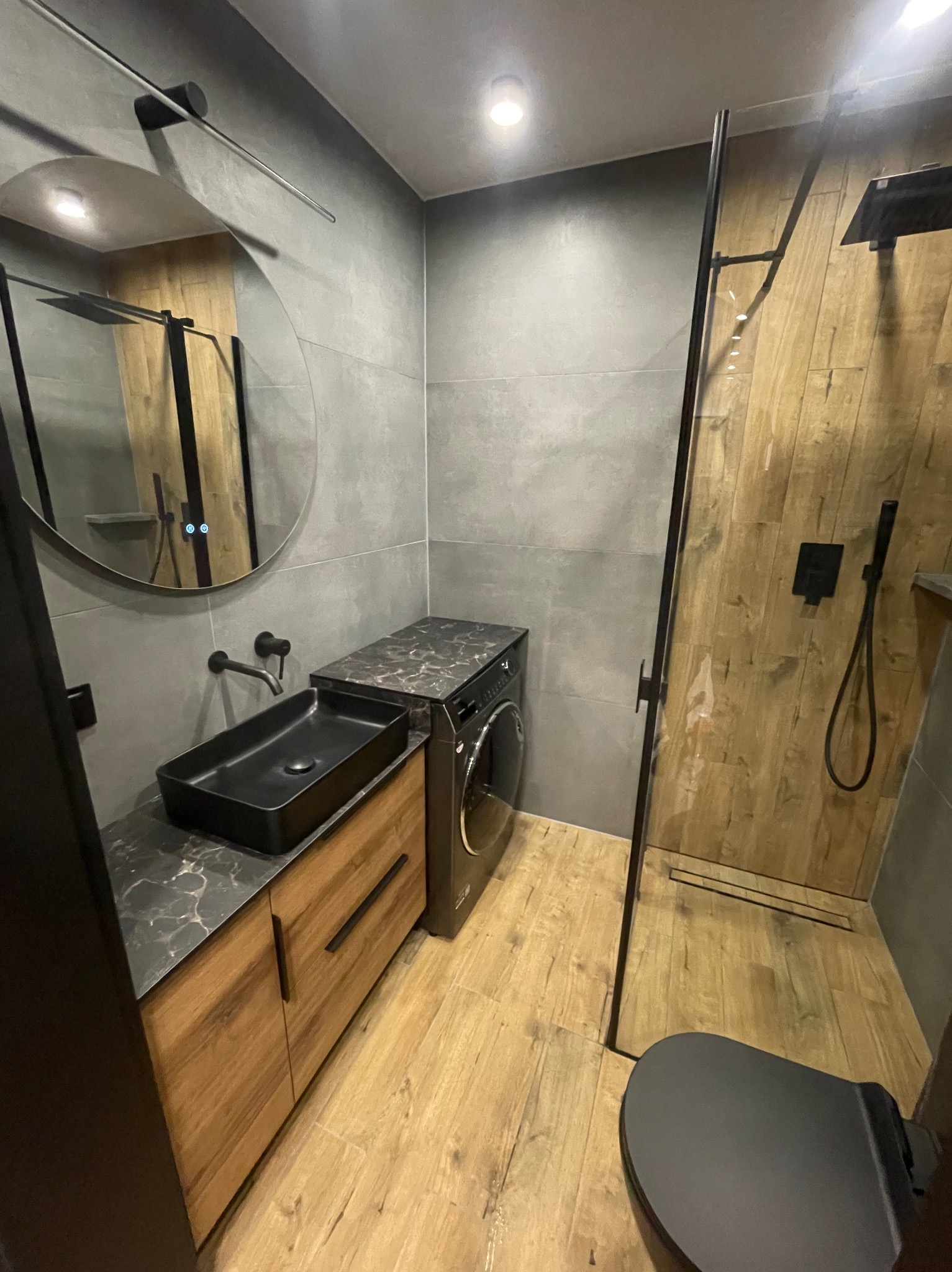 Nowoczesna łazienka z kabiną prysznicową typu walk-in, umywalką nablatową i pralką pod blatem. Minimalistyczny design z drewnem i szarością.