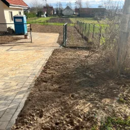 Ułożona kostka brukowa przy ogrodzeniu i wyrównany teren pod trawnik, w tle widoczny dom i budynki gospodarcze.