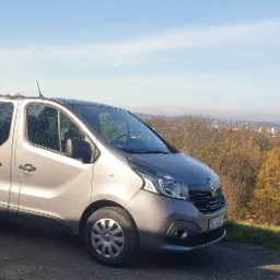 Szary van marki Renault na tle jesiennego krajobrazu z kominem i kościołem w oddali.