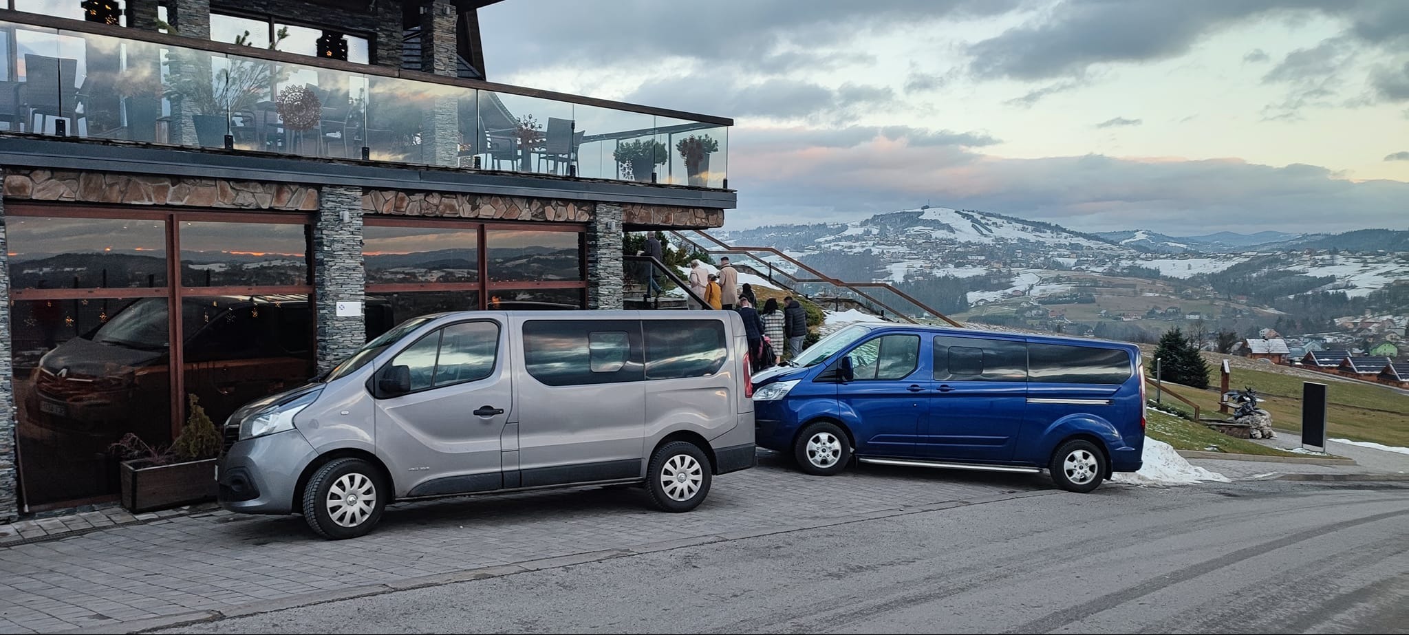 Dwa busy, szary Renault Trafic i niebieski Ford Transit, zaparkowane przed budynkiem z kamienną elewacją na tle górskiego krajobrazu z widocznymi zabudowaniami i śniegiem.