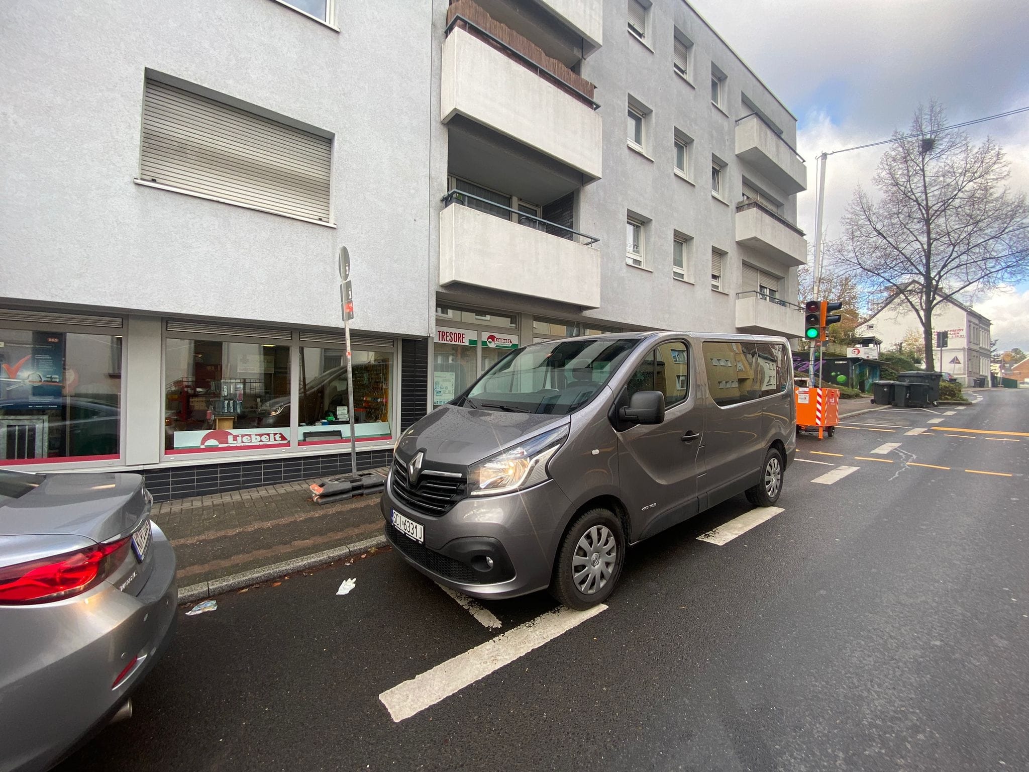 Szary van Renault zaparkowany na mokrej ulicy przed budynkiem mieszkalnym z balkonami, w tle sygnalizacja świetlna z zielonym światłem.