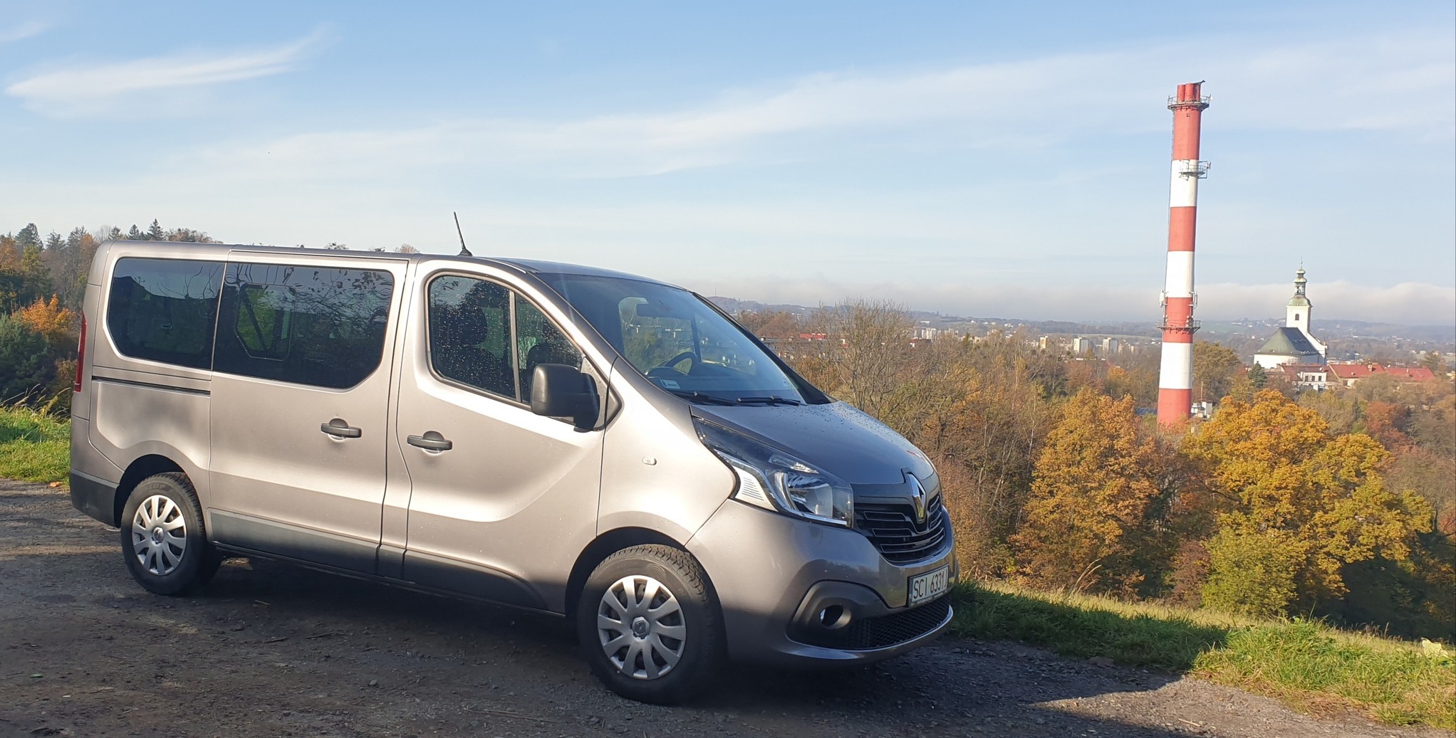 Szary van marki Renault na tle jesiennego krajobrazu z kominem i kościołem w oddali.